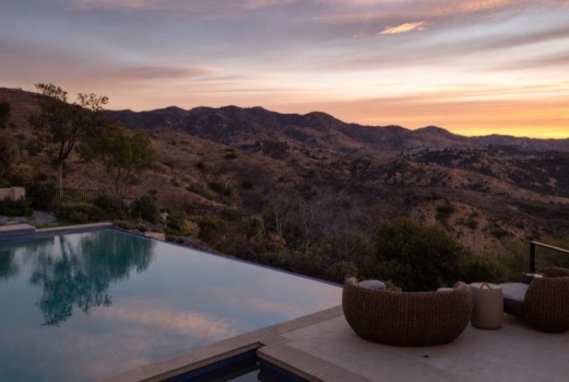 Pool deck of a modern house set into a slope, with layered hills turning gold and green at dusk